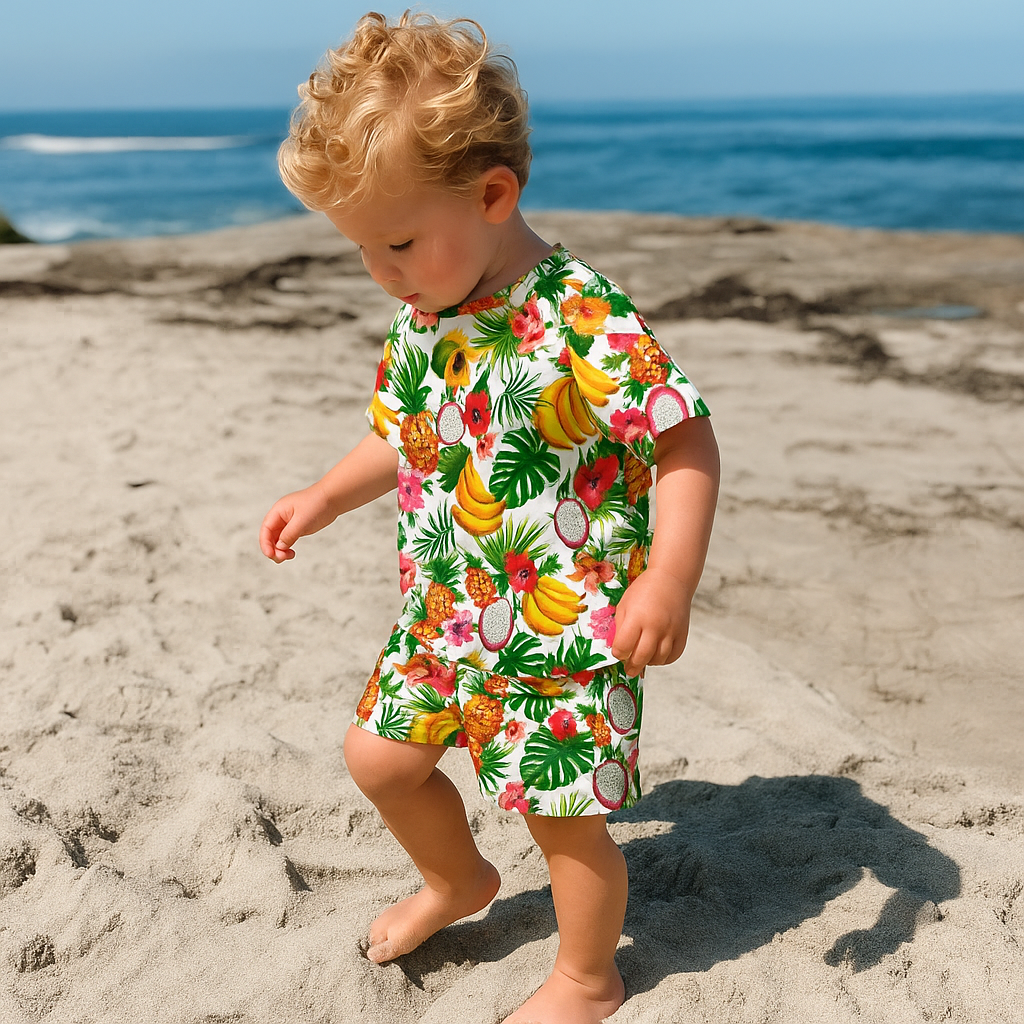 Child wearing a colorful tropical fruit patterned outfit on a sandy beach with ocean in the background