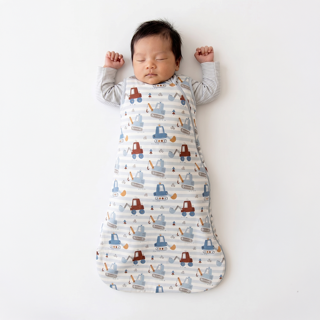 A sleeping newborn baby lying on their back, swaddled in a long, soft baby blanket or sleep sack with a playful construction vehicle pattern. The blanket features cartoon trucks, excavators, and cranes in blue, red, and orange on a white background with light gray stripes.