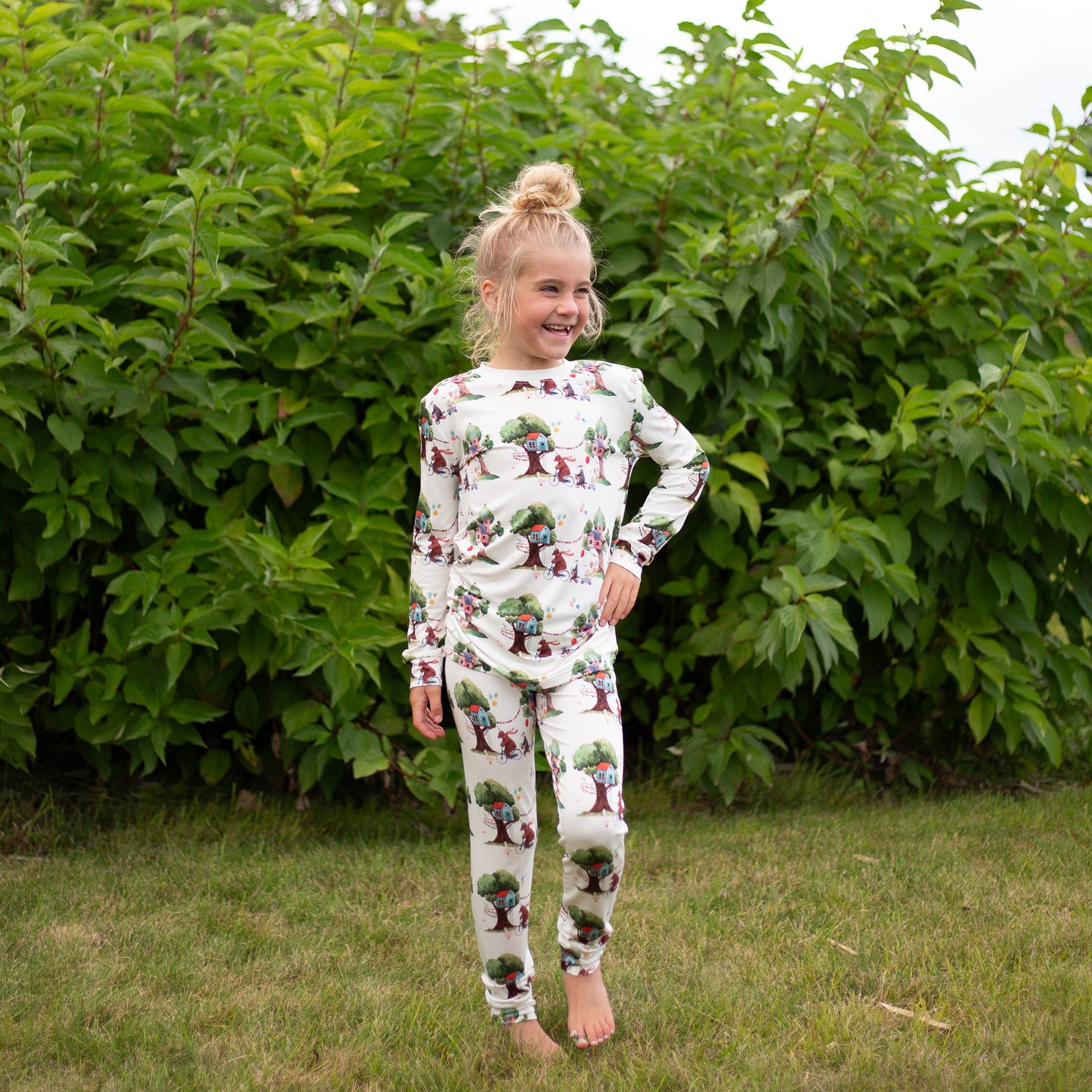 Young girl wearing a long-sleeve pajama set with a nature print, standing in front of green bushes.