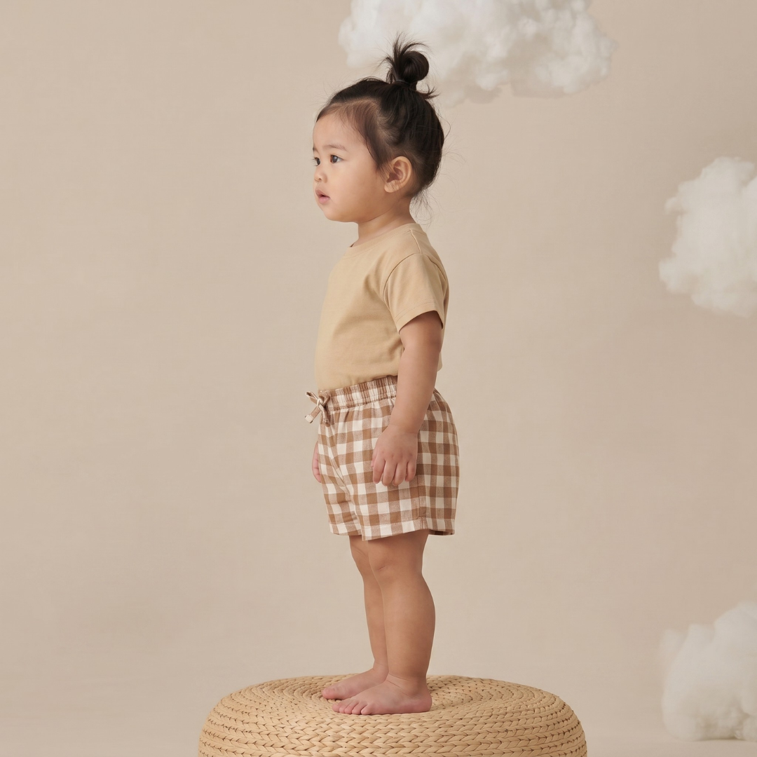 Child wearing a beige shirt and checkered shorts standing on a woven stool against a beige background with clouds.