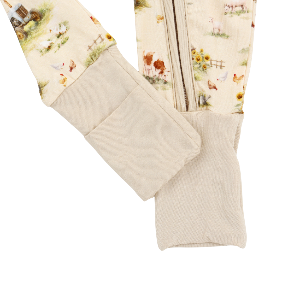 Close-up of a cream-colored fleece footed sleeper with a printed back panel featuring a farm scene with chickens, cows, and sunflowers, and a zippered closure.