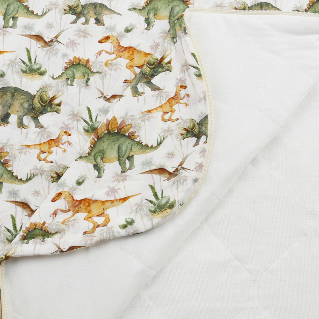 Close-up of a quilted baby blanket showing a dinosaur print side with green and orange dinosaurs on a white background, and a plain white quilted reverse side.