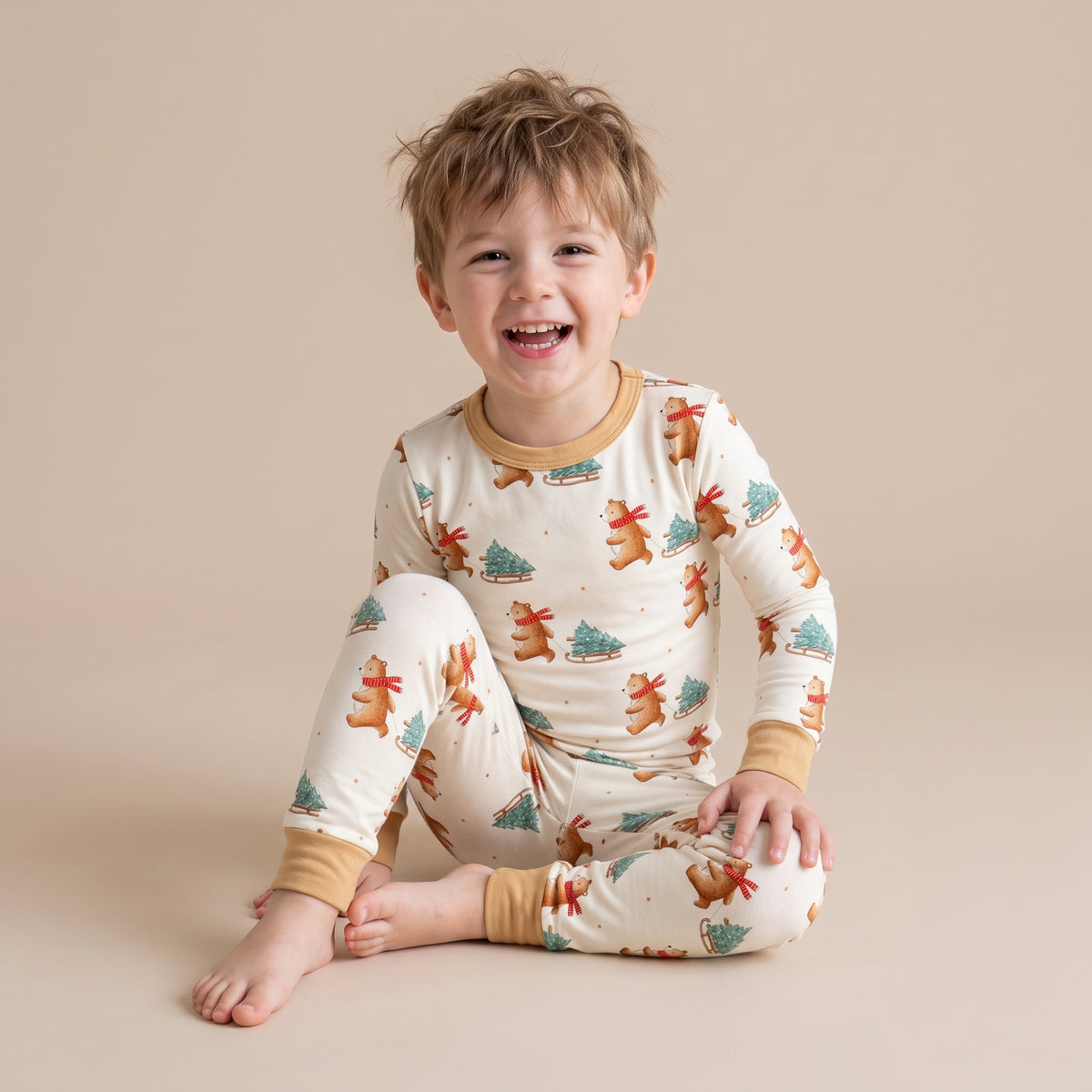 A young child sitting on a plain beige studio background, wearing a cream-colored long-sleeve pajama set with a holiday print of gingerbread cookies, Christmas trees, and ornaments, with tan ribbed cuffs and collar.