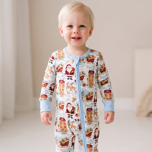 A smiling toddler wearing a long-sleeve zip-front pajama romper with a light blue trim and a festive holiday print featuring Santa Claus, snowmobiles, houses, and deer on a cream background.
