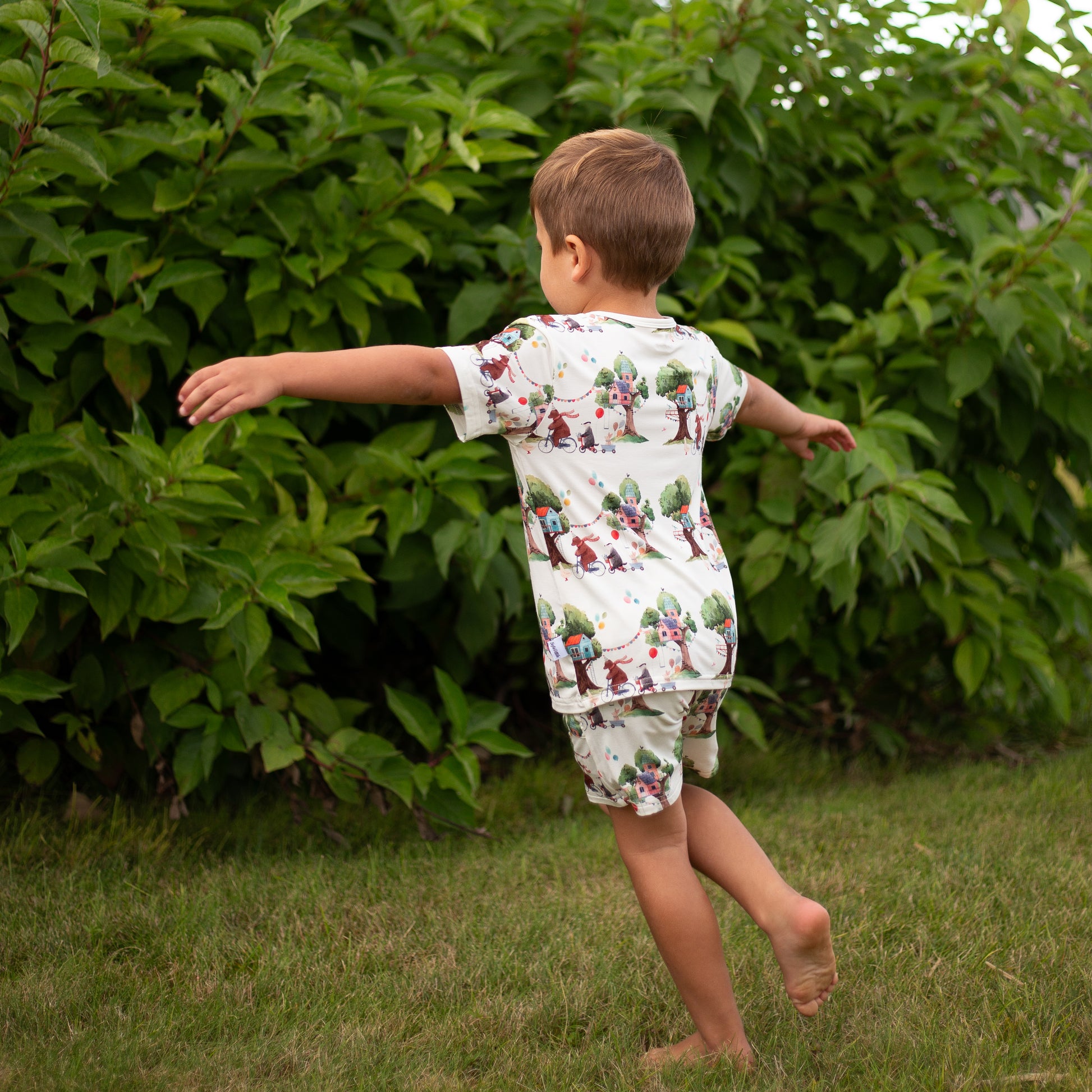 Child in a patterned outfit running in a grassy area with green bushes.